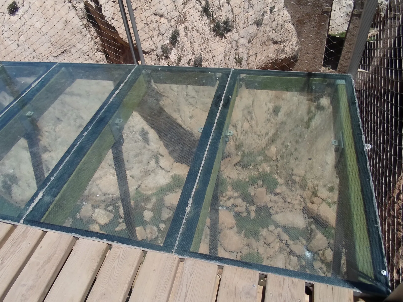 A short glass-floor viewing balcony on the Caminito del Rey boardwalk, looking straight down through transparent panels to the rocky gorge floor far below